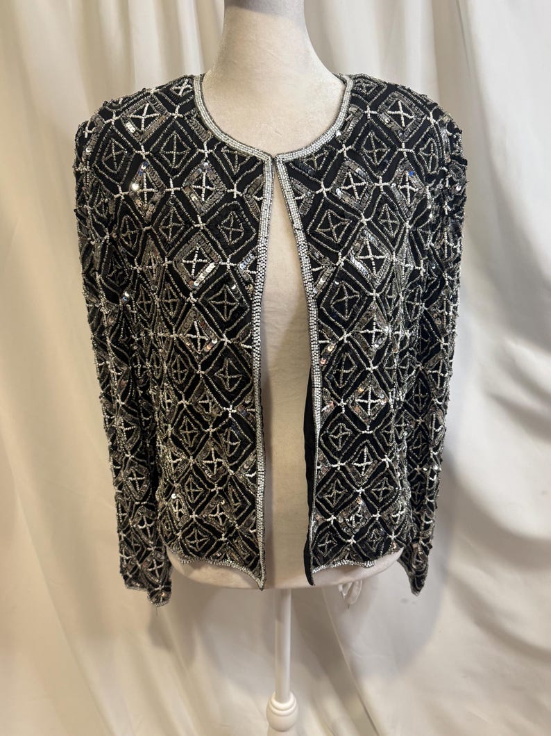 May include: A black open-front jacket featuring an intricate geometric pattern created with silver beads and sequins. The garment has long sleeves, a round neckline, and silver beaded trim along the edges, showcasing a dense, textured surface.