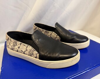 May include: A pair of slip-on sneakers featuring a smooth black leather front panel and a textured grey and black snakeskin pattern on the sides and heel. The shoes have a thick white rubber sole and a small beige tab at the back of the heel.