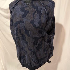K&ouml;nnte beinhalten: Ein lang&auml;rmeliger Pullover mit einem dunkelblauen und anthrazitfarbenen Tarnmuster. Das Kleidungsst&uuml;ck hat einen gerippten Rundhalsausschnitt und gerippte B&uuml;ndchen am Saum. Ein schwarzes Etikett am inneren Halsausschnitt zeigt den Text Barefoot Dreams.