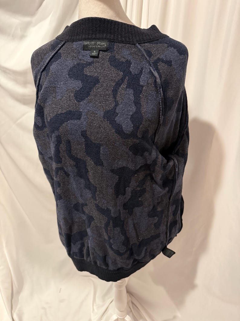 K&ouml;nnte beinhalten: Ein lang&auml;rmeliger Pullover mit einem dunkelblauen und anthrazitfarbenen Tarnmuster. Das Kleidungsst&uuml;ck hat einen gerippten Rundhalsausschnitt und gerippte B&uuml;ndchen am Saum. Ein schwarzes Etikett am inneren Halsausschnitt zeigt den Text Barefoot Dreams.