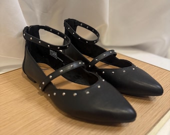 May include: A pair of black flats with pointed toes and ankle straps. The flats have silver stud accents on the straps and around the ankle. The shoes are displayed on a light wood surface.