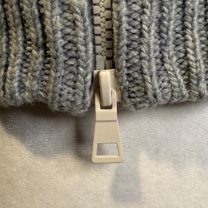 Puede incluir: Primer plano de una tela de punto gris con una cremallera blanca. La tela tiene una textura acanalada. El tirador de la cremallera es de color beige claro.