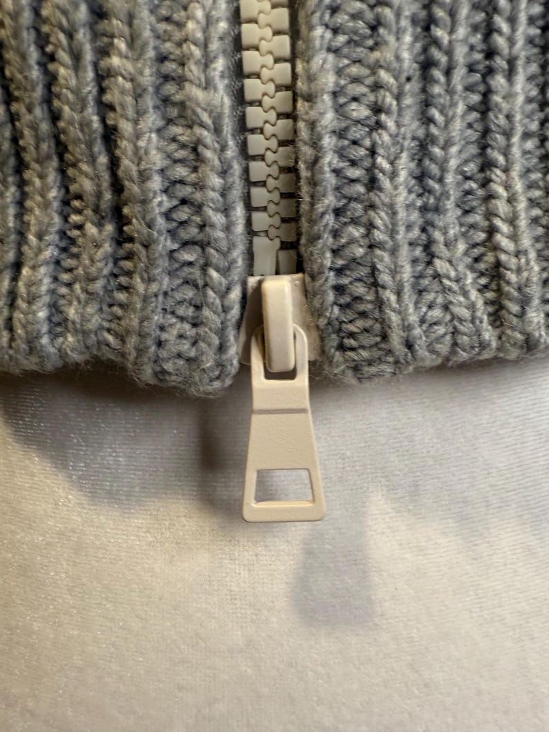 Puede incluir: Primer plano de una tela de punto gris con una cremallera blanca. La tela tiene una textura acanalada. El tirador de la cremallera es de color beige claro.