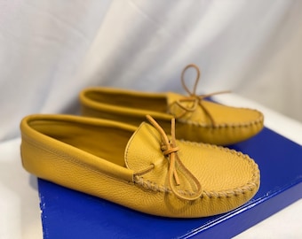 May include: A pair of mustard yellow leather moccasins featuring a textured surface, a rounded toe, and a decorative lace-up tie detail on the top. The shoes have visible whipstitch detailing along the perimeter of the toe box and a slip-on design.