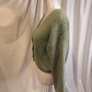 May include: A sage green, cropped cardigan sweater with a V-neck and long sleeves. The sweater has a textured, nubby knit and is buttoned up the front. The garment is displayed on a mannequin against a white backdrop.