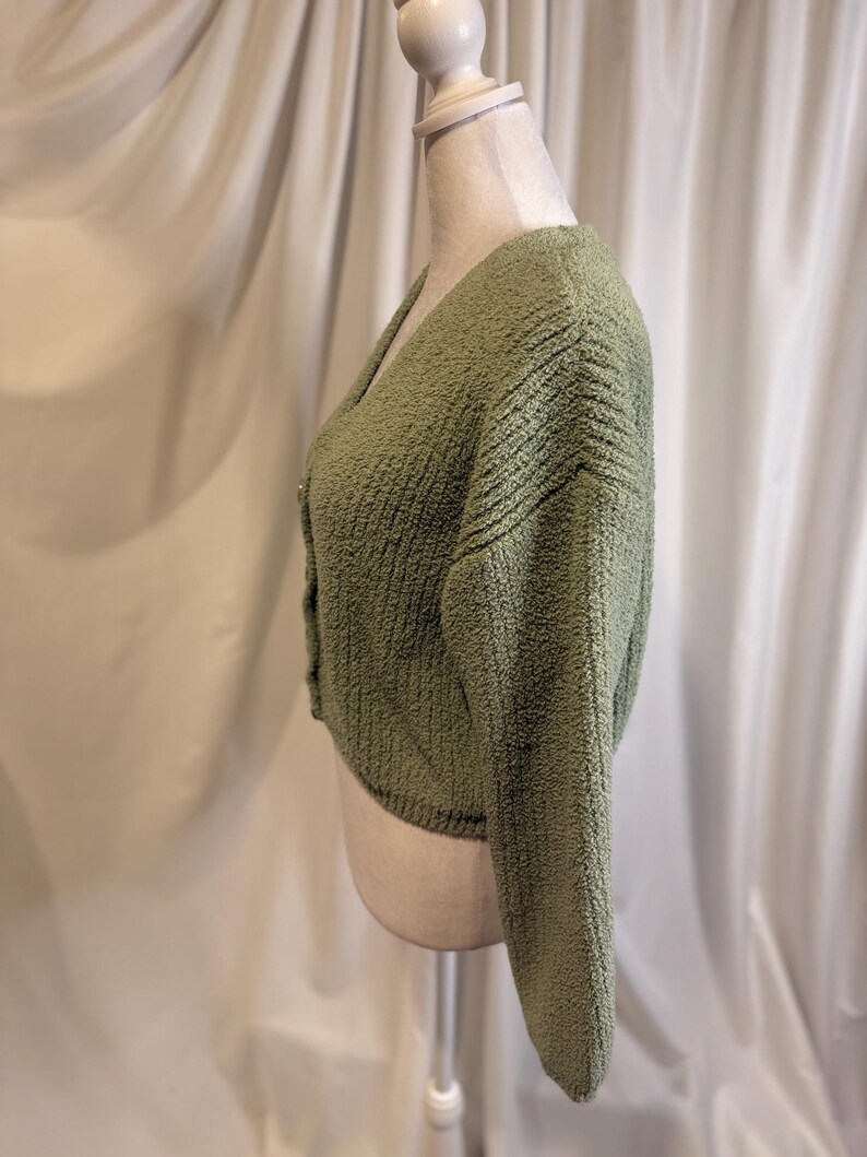 May include: A sage green, cropped cardigan sweater with a V-neck and long sleeves. The sweater has a textured, nubby knit and is buttoned up the front. The garment is displayed on a mannequin against a white backdrop.