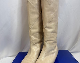 May include: A pair of knee-high boots in a light cream color, crafted with a smooth finish. The boots feature a distinct diamond-shaped quilted pattern across the shaft, a pointed toe design, and a flat sole, presenting a clean and structured look.