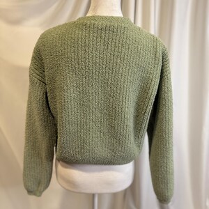 May include: A sage green, textured knit sweater. The sweater has a crew neck and long sleeves. The back of the sweater is visible, showing the texture of the knit fabric. The sweater is displayed on a mannequin.