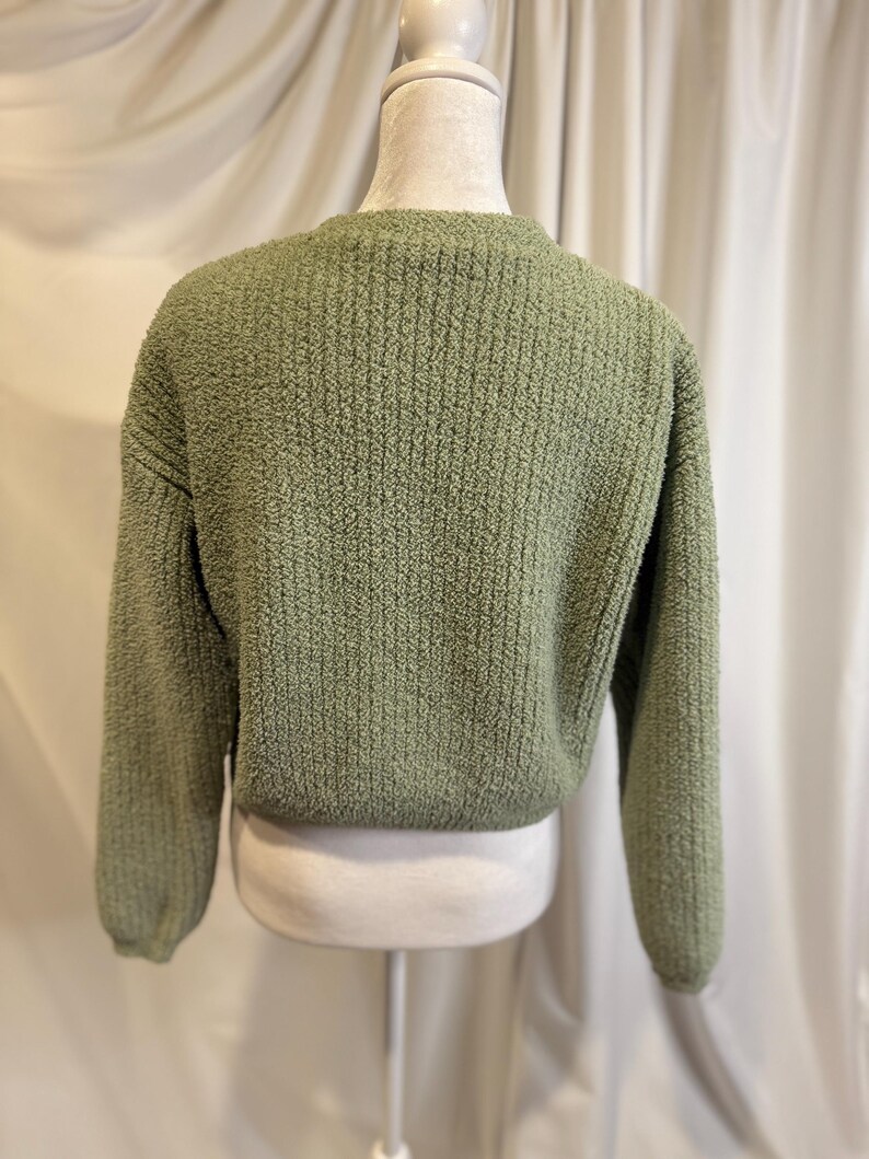 May include: A sage green, textured knit sweater. The sweater has a crew neck and long sleeves. The back of the sweater is visible, showing the texture of the knit fabric. The sweater is displayed on a mannequin.