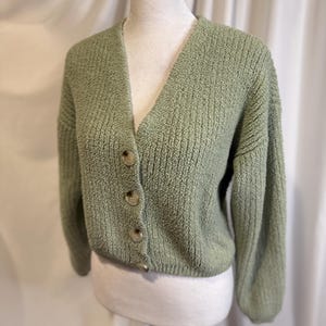 May include: A sage green cardigan sweater with a V-neck and three large, round buttons. The sweater has a textured knit design and long sleeves. The garment is displayed on a mannequin.