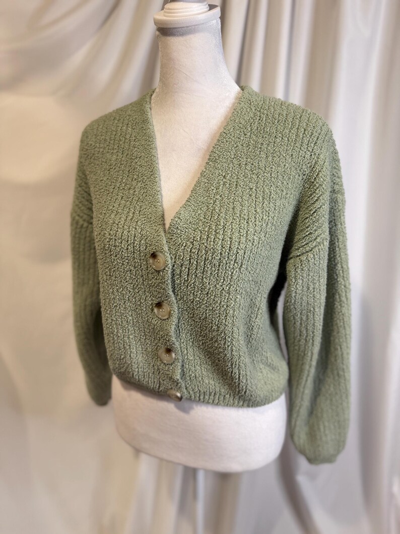 May include: A sage green cardigan sweater with a V-neck and three large, round buttons. The sweater has a textured knit design and long sleeves. The garment is displayed on a mannequin.