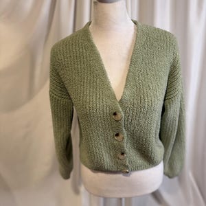 May include: A sage green cardigan with a V-neck and long sleeves. The textured knit sweater has four round, light brown buttons down the front. The cardigan is displayed on a white mannequin.