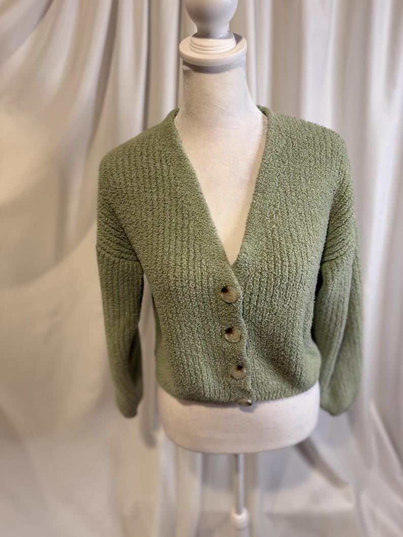 May include: A sage green cardigan with a V-neck and long sleeves. The textured knit sweater has four round, light brown buttons down the front. The cardigan is displayed on a white mannequin.