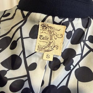 Puede incluir: Primer plano de un cuello de abrigo azul marino y el forro interior. El forro presenta un patr&oacute;n abstracto en blanco y negro. Una etiqueta rectangular con el texto "Tulle original clothing" est&aacute; cosida en el forro, con una talla "S" debajo.
