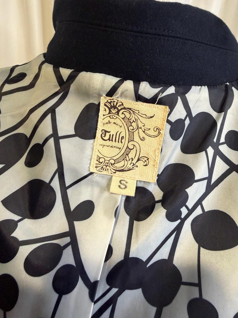 Puede incluir: Primer plano de un cuello de abrigo azul marino y el forro interior. El forro presenta un patr&oacute;n abstracto en blanco y negro. Una etiqueta rectangular con el texto "Tulle original clothing" est&aacute; cosida en el forro, con una talla "S" debajo.