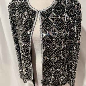 May include: A black open-front jacket featuring an intricate geometric pattern created with silver sequins and white beads. The garment has a round neckline and long sleeves, with the entire surface covered in a dense, repeating diamond-shaped design.