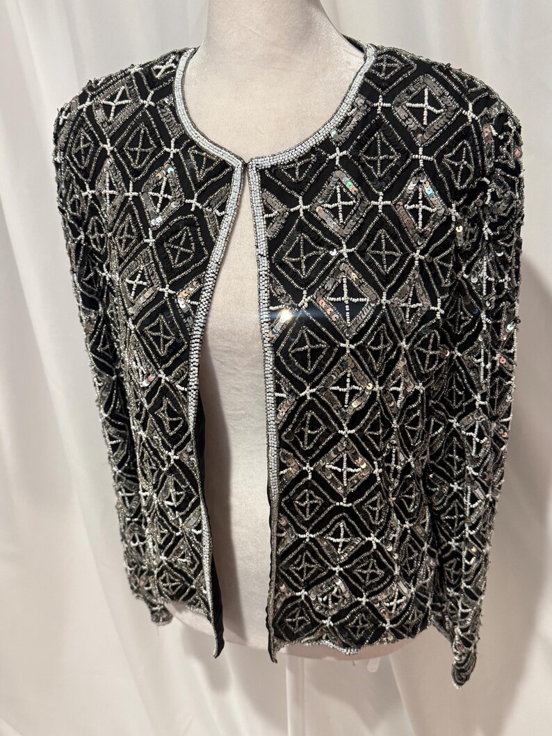 May include: A black open-front jacket featuring an intricate geometric pattern created with silver sequins and white beads. The garment has a round neckline and long sleeves, with the entire surface covered in a dense, repeating diamond-shaped design.