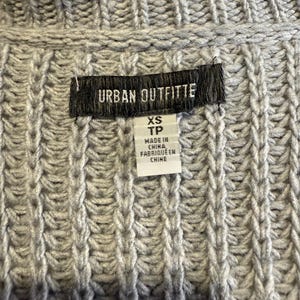 Puede incluir: Primer plano de un su&eacute;ter de punto gris con textura acanalada. La etiqueta dice "URBAN OUTFITTERS" e incluye la talla "XS TP" y "MADE IN CHINA / FABRIQUE EN CHINE". El su&eacute;ter est&aacute; hecho de un punto grueso y texturizado.