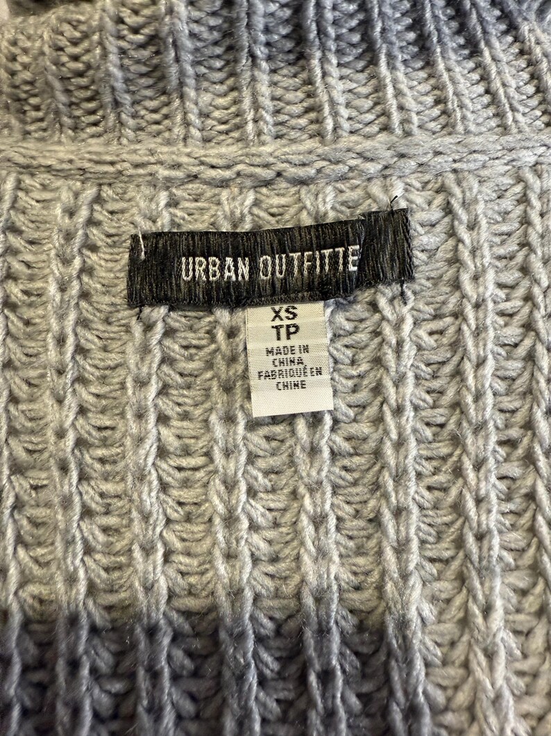 Puede incluir: Primer plano de un su&eacute;ter de punto gris con textura acanalada. La etiqueta dice "URBAN OUTFITTERS" e incluye la talla "XS TP" y "MADE IN CHINA / FABRIQUE EN CHINE". El su&eacute;ter est&aacute; hecho de un punto grueso y texturizado.