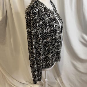 May include: A long-sleeved jacket featuring a black base fabric adorned with an intricate geometric pattern of silver and white beads. The garment has a round neckline, a straight hem, and a front opening, showcasing dense beadwork throughout.