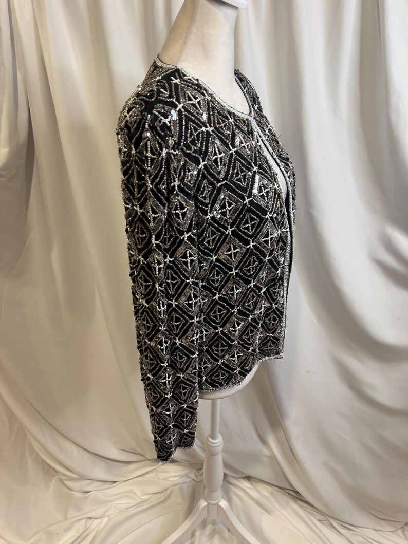 May include: A long-sleeved jacket featuring a black base fabric adorned with an intricate geometric pattern of silver and white beads. The garment has a round neckline, a straight hem, and a front opening, showcasing dense beadwork throughout.