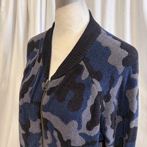 K&ouml;nnte beinhalten: Eine Bomberjacke mit Rei&szlig;verschluss und Camouflage-Muster in Marineblau, Anthrazit und Schwarz. Die Jacke hat einen gerippten schwarzen Kragen und einen Metallrei&szlig;verschluss auf der Vorderseite, pr&auml;sentiert auf einem Schaufensterpuppen-Torso.