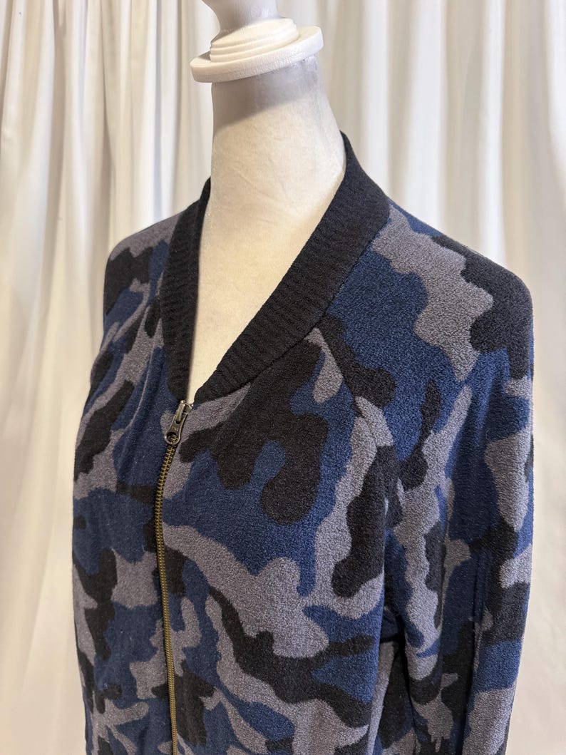 K&ouml;nnte beinhalten: Eine Bomberjacke mit Rei&szlig;verschluss und Camouflage-Muster in Marineblau, Anthrazit und Schwarz. Die Jacke hat einen gerippten schwarzen Kragen und einen Metallrei&szlig;verschluss auf der Vorderseite, pr&auml;sentiert auf einem Schaufensterpuppen-Torso.