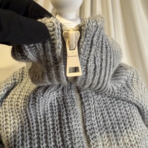 Puede incluir: Primer plano de un su&eacute;ter de punto gris con cuello alto y cremallera color crema. El su&eacute;ter tiene una textura de punto grueso y un cuello acanalado. La cremallera est&aacute; completamente cerrada y el su&eacute;ter parece estar hecho de un material suave y c&aacute;lido.