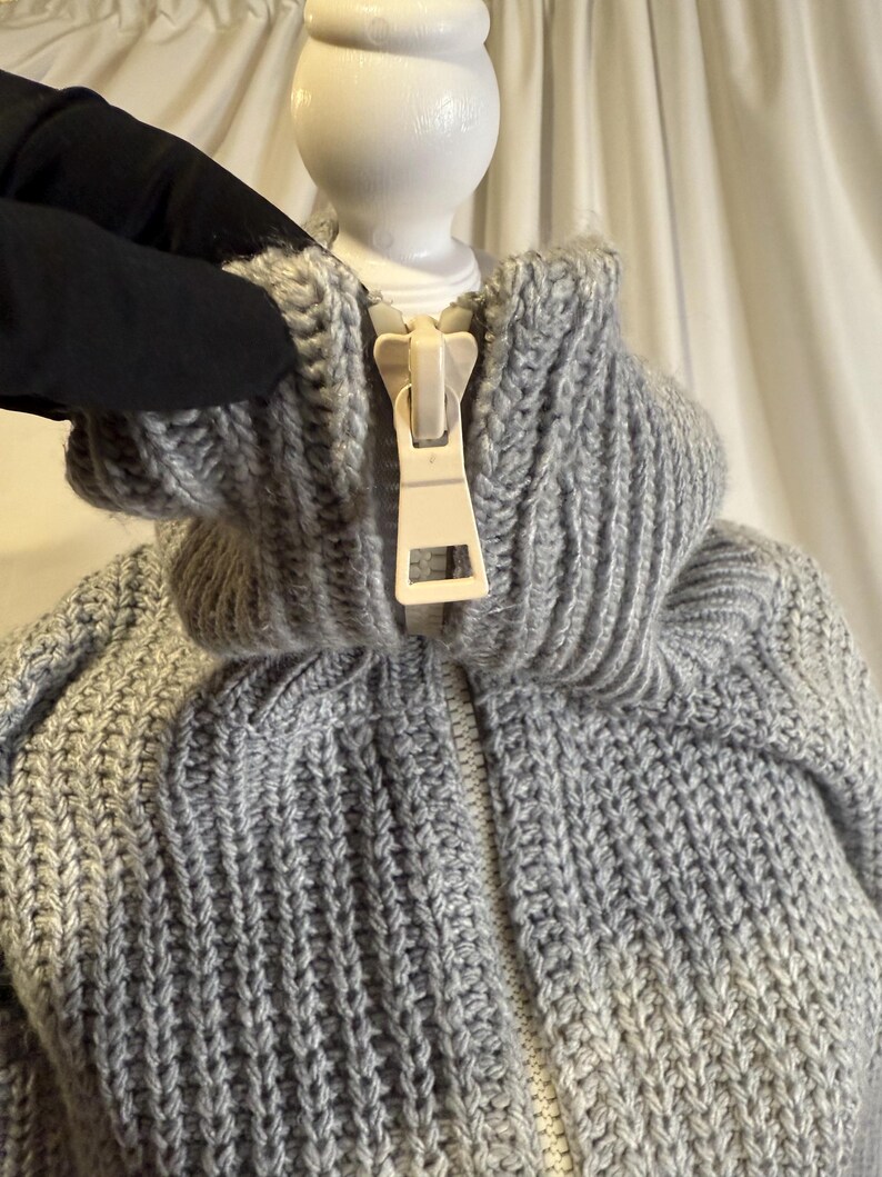 Puede incluir: Primer plano de un su&eacute;ter de punto gris con cuello alto y cremallera color crema. El su&eacute;ter tiene una textura de punto grueso y un cuello acanalado. La cremallera est&aacute; completamente cerrada y el su&eacute;ter parece estar hecho de un material suave y c&aacute;lido.