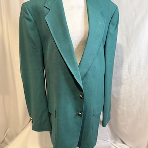 May include: A green blazer with gold-colored buttons and two front pockets. The blazer is displayed on a white mannequin. The fabric has a subtle texture. The blazer has a classic, tailored design.