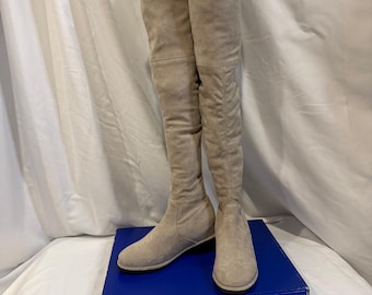 May include: A pair of beige over-the-knee boots. The boots are made of a soft material and have a low heel. They are displayed on a blue box against a white backdrop.