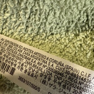 May include: Close-up of a light green, textured fabric. A white label with black text is partially visible, displaying text including company information and product details. The fabric has a soft, plush appearance.