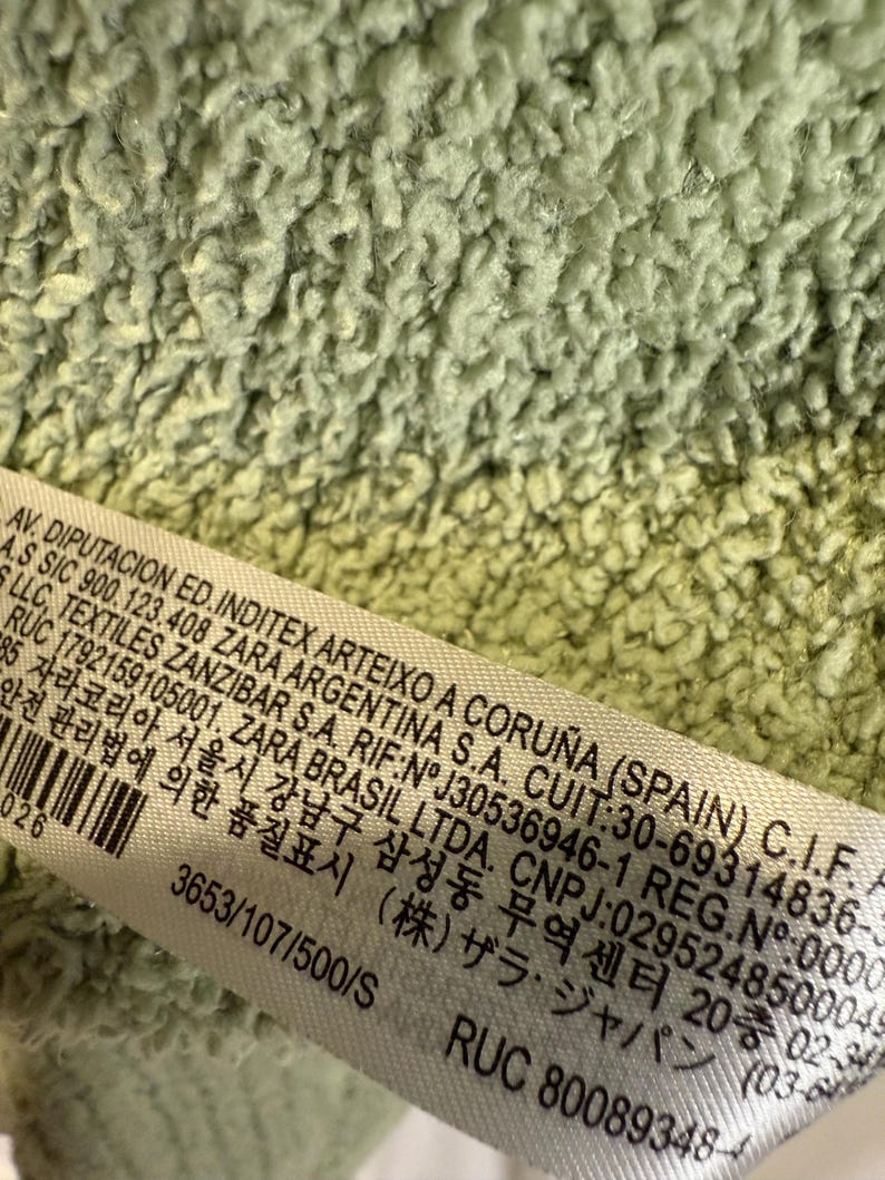 May include: Close-up of a light green, textured fabric. A white label with black text is partially visible, displaying text including company information and product details. The fabric has a soft, plush appearance.