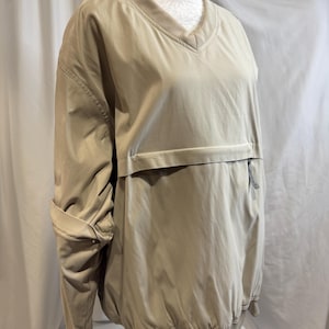 May include: A tan pullover jacket with a V-neck and a front pocket. The jacket has long sleeves that are rolled up. The bottom hem and cuffs are elasticized. The jacket is displayed on a mannequin.