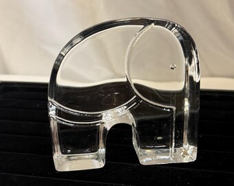 May include: A clear glass elephant figurine. The elephant has a curved back, a large ear, and a small trunk. The figurine is transparent and sits on a black surface. The elephant's form is simple and elegant.