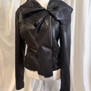 May include: A black jacket with a unique design. The jacket features a large bow detail on the shoulder and a zipper closure. The material has a textured, metallic appearance. The jacket is displayed on a mannequin.
