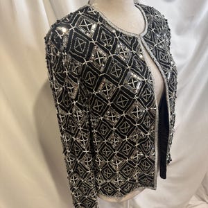 May include: A black long-sleeve jacket featuring an intricate geometric pattern created with silver sequins and white beads. The open-front garment has a round neckline and is fully embellished with a repeating diamond grid design throughout.