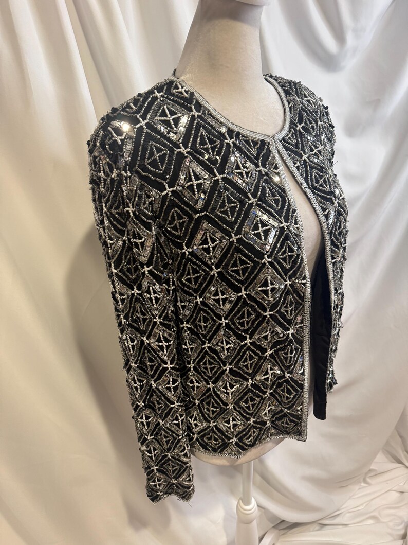 May include: A black long-sleeve jacket featuring an intricate geometric pattern created with silver sequins and white beads. The open-front garment has a round neckline and is fully embellished with a repeating diamond grid design throughout.