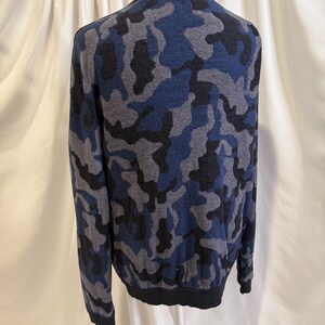 K&ouml;nnte beinhalten: Ein lang&auml;rmeliger Strickpullover mit Camouflage-Muster in Marineblau, Anthrazit und Schwarz. Das Kleidungsst&uuml;ck hat einen einfarbigen schwarzen gerippten Kragen, B&uuml;ndchen und Saum und wird auf einer Schaufensterpuppe pr&auml;sentiert, um die Form zu zeigen.