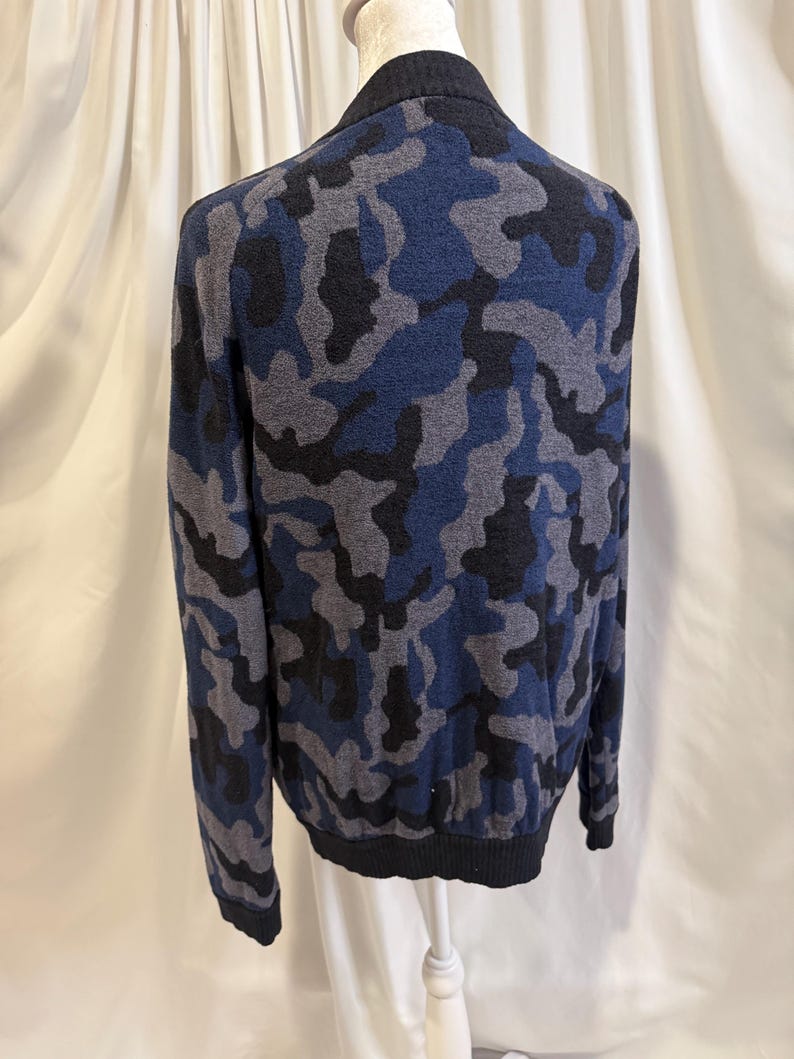 K&ouml;nnte beinhalten: Ein lang&auml;rmeliger Strickpullover mit Camouflage-Muster in Marineblau, Anthrazit und Schwarz. Das Kleidungsst&uuml;ck hat einen einfarbigen schwarzen gerippten Kragen, B&uuml;ndchen und Saum und wird auf einer Schaufensterpuppe pr&auml;sentiert, um die Form zu zeigen.