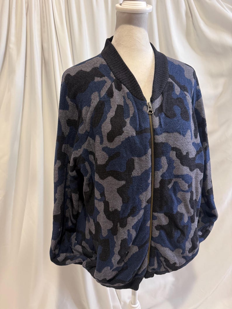 K&ouml;nnte beinhalten: Eine lang&auml;rmelige Bomberjacke mit Camouflage-Muster in Marineblau, Anthrazit und Schwarz. Die Jacke verf&uuml;gt &uuml;ber einen gerippten schwarzen Kragen, B&uuml;ndchen und Saum sowie einen durchgehenden Metallrei&szlig;verschluss an der Vorderseite.