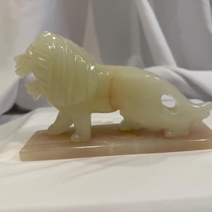 May include: A white jade lion sculpture on a pink marble base. The lion is standing on all four paws with its head turned to the side. The sculpture is detailed and intricate.