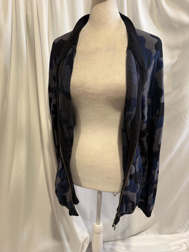 K&ouml;nnte beinhalten: Eine lang&auml;rmelige Jacke mit Rei&szlig;verschluss und Camouflage-Muster in Marineblau, Anthrazit und Schwarz. Das Kleidungsst&uuml;ck hat einen einfarbigen, schwarzen Rippkragen und passende Rippb&uuml;ndchen sowie einen durchgehenden Metallrei&szlig;verschluss.