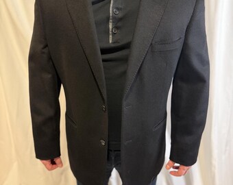 May include: A black blazer with a two-button closure, paired with a black Henley shirt and dark blue jeans. The blazer has a classic design with a notched lapel and a pocket on the left side.