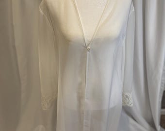 May include: A sheer white robe with long sleeves and a V-neckline. The robe features delicate lace trim at the cuffs and hem. A single button closure is at the waist. The robe is displayed on a mannequin.