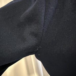 Puede incluir: Primer plano de una prenda azul marino oscuro, posiblemente un abrigo o una chaqueta, que muestra el hombro y la manga. La tela parece ser lana texturizada o un material similar. La costura del hombro es visible.