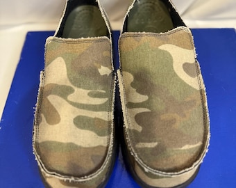 May include: A pair of slip-on shoes featuring a green, brown, and tan camouflage pattern on the canvas upper. The shoes have a distinct frayed edge detail along the seams, a rounded toe box, and a dark interior lining visible at the opening.