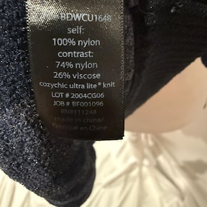 K&ouml;nnte beinhalten: Ein schwarzes Pflegeetikett mit wei&szlig;em Text: BDWCU1648, self 100% nylon, contrast 74% nylon und 26% viscose, cozychic ultra lite knit, LOT 2004CG06, JOB BF001096, RN 111248 sowie made in china, befestigt an einem dunkelblauen, strukturierten Strickstoff.