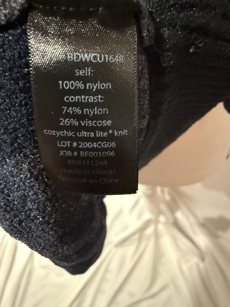 K&ouml;nnte beinhalten: Ein schwarzes Pflegeetikett mit wei&szlig;em Text: BDWCU1648, self 100% nylon, contrast 74% nylon und 26% viscose, cozychic ultra lite knit, LOT 2004CG06, JOB BF001096, RN 111248 sowie made in china, befestigt an einem dunkelblauen, strukturierten Strickstoff.
