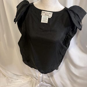 May include: A black sleeveless top with structured, pleated shoulder details and a round neckline. The interior label reads Papell Boutique Evening, size M. The garment features a solid black fabric construction with a gathered hemline at the waist.